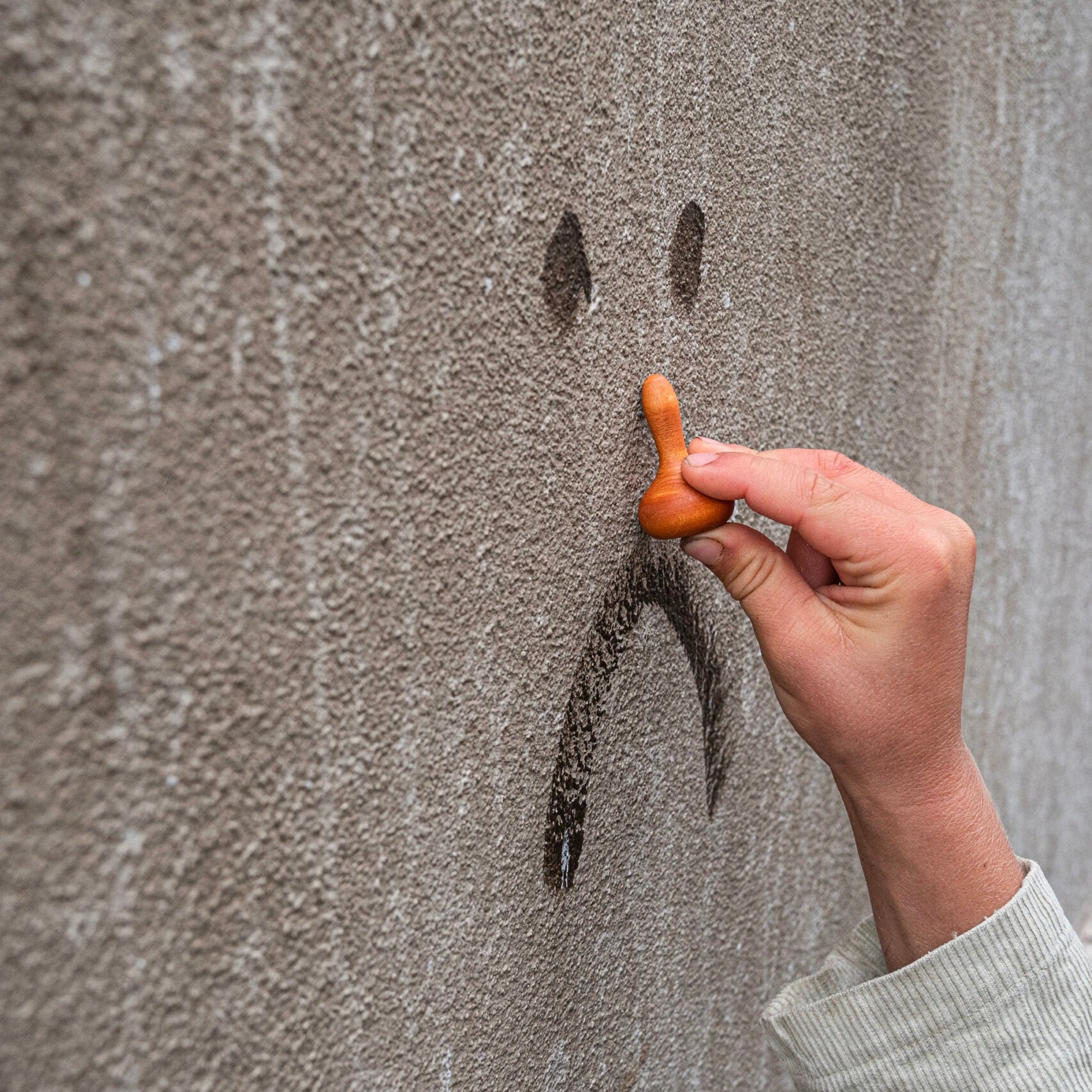 Hand holding a wooden spoon above a smiley face drawn on a textured wall.