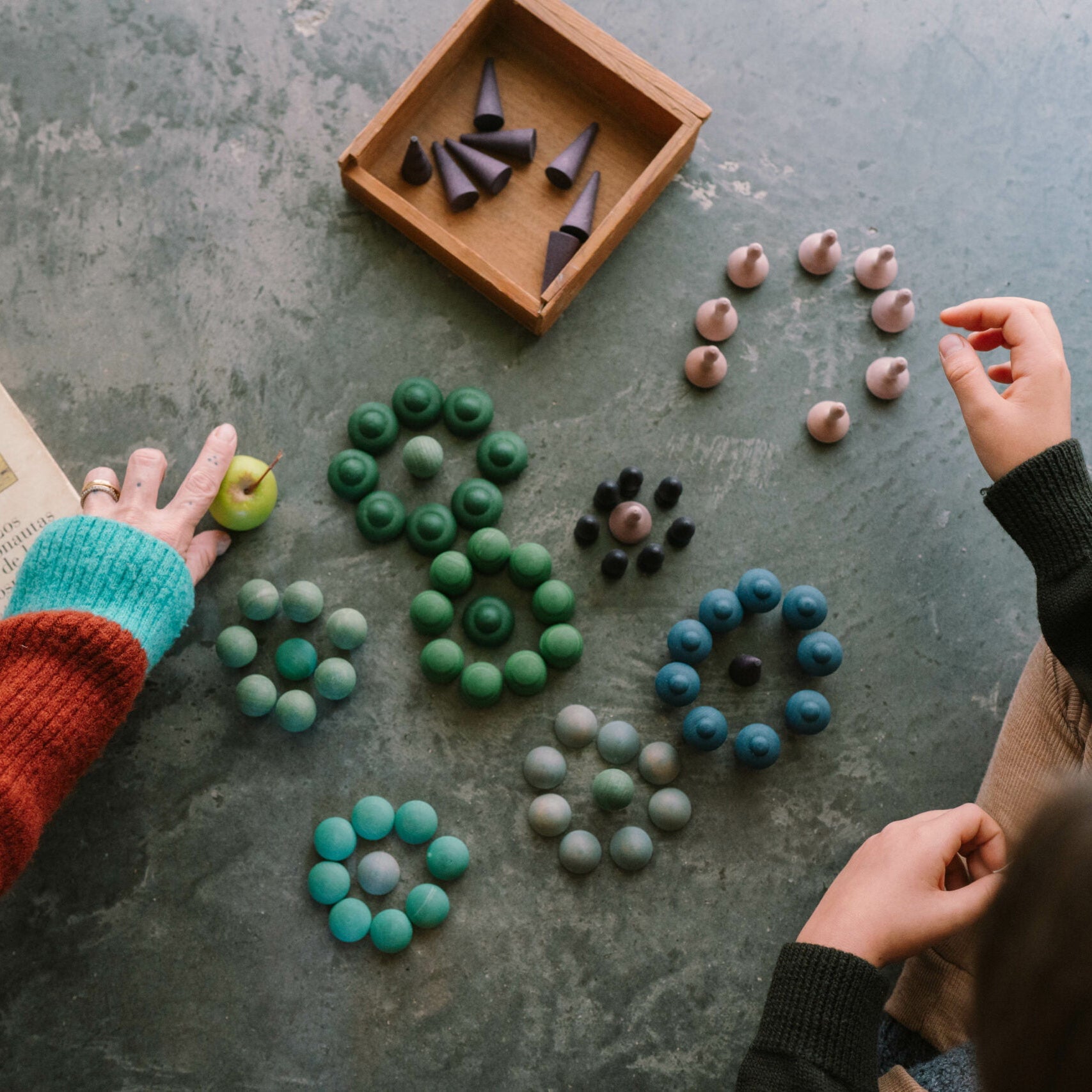 Two people arranging colorful beads on a textured surface with a book and wooden box.