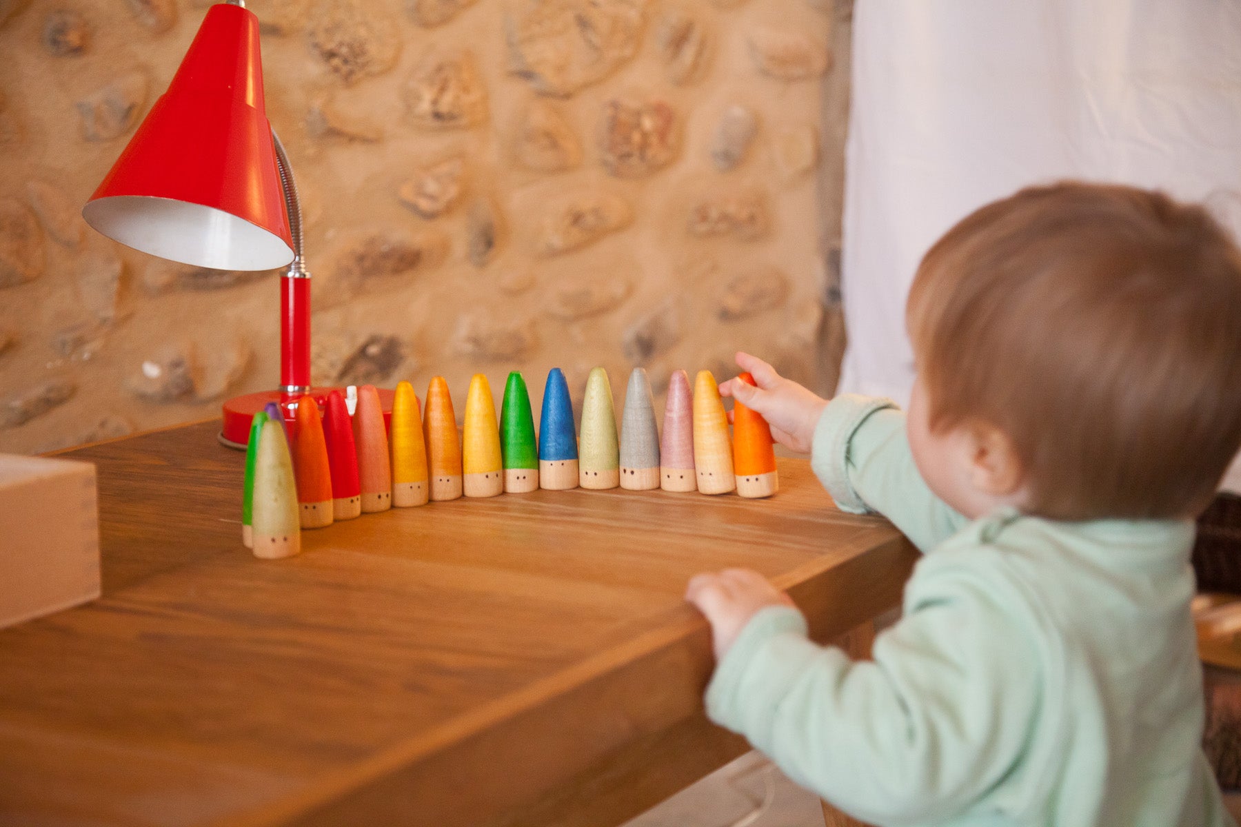 Child playing with colorful crayons on a wooden surface next to a red lamp.