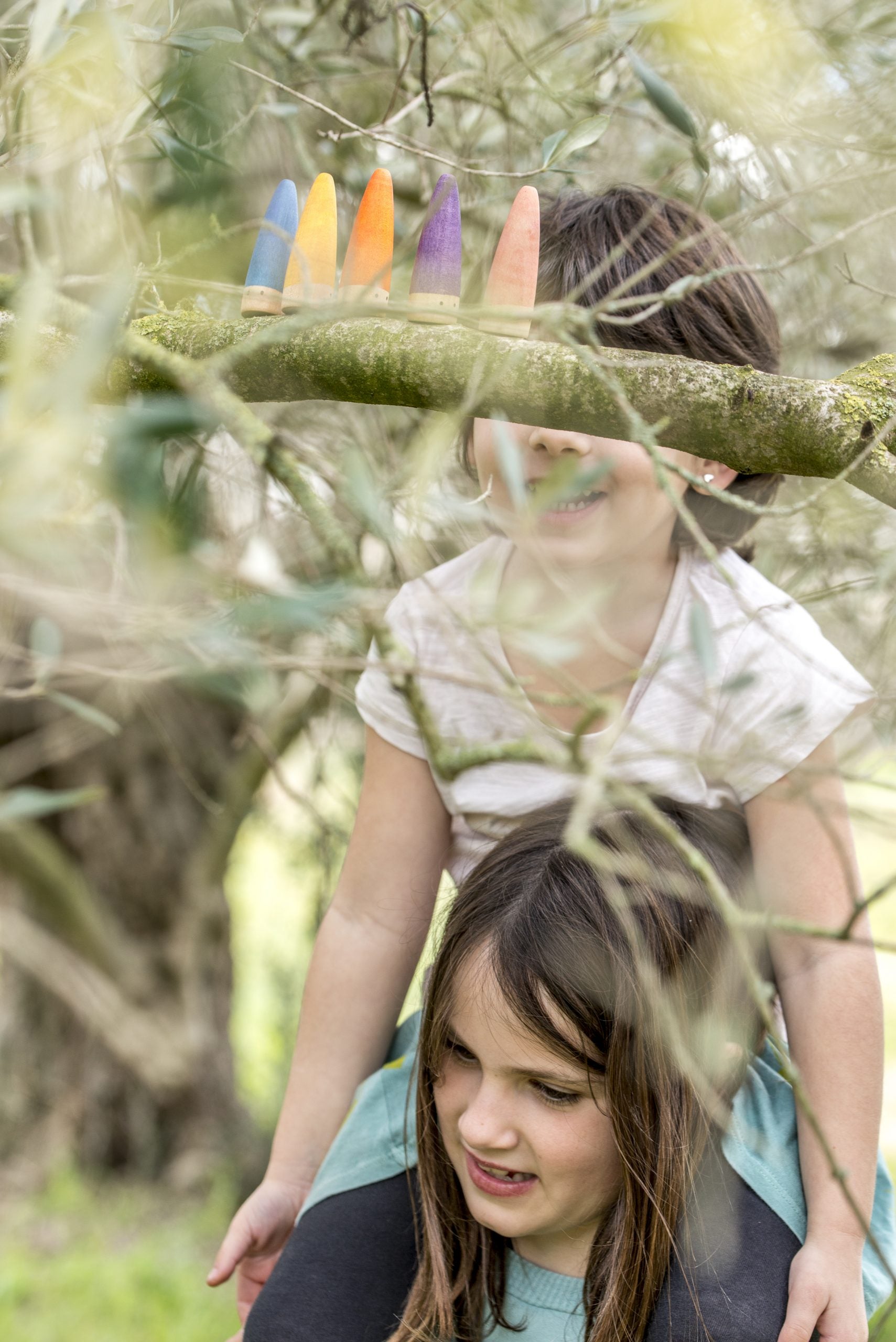 Two children playing outdoors with colorful sticks, surrounded by greenery.