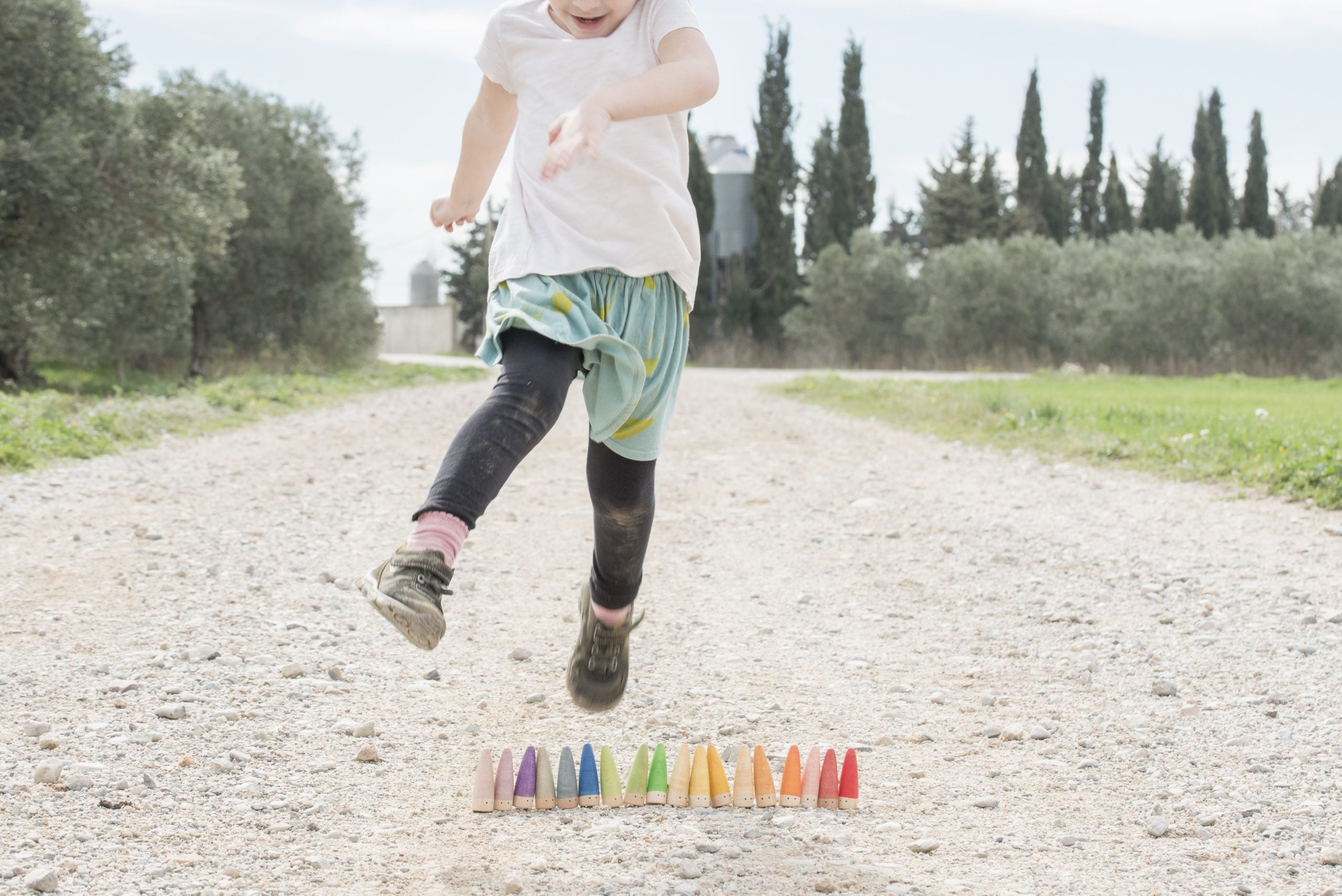 Child jumping over colorful sticks on a gravel path with trees in the background