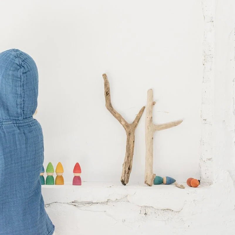 Child in a blue hooded outfit standing in front of a white wall with colorful toys and wooden sticks.