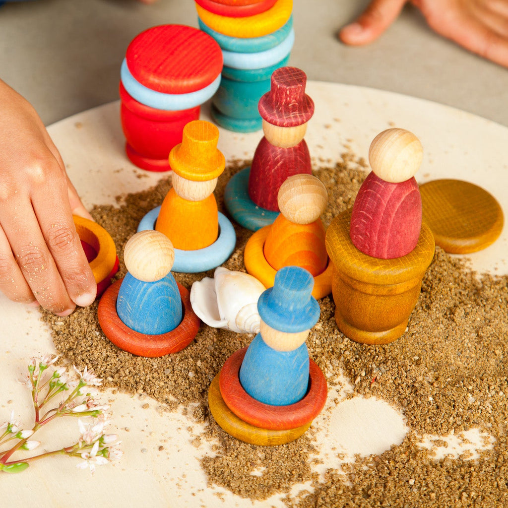 Colorful wooden toy figures on a sand surface with a child's hand interacting with them.