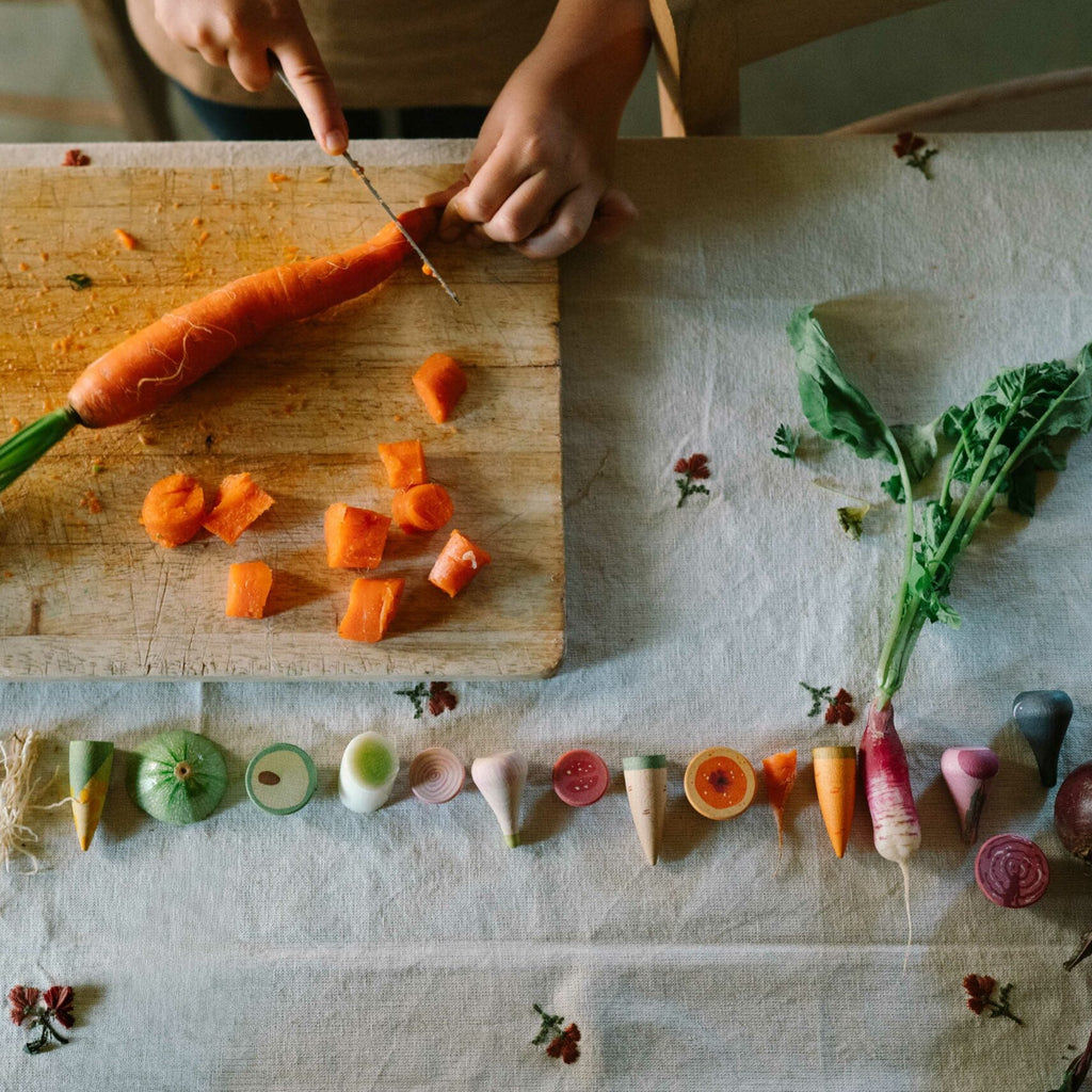 Person cutting a carrot on a wooden cutting board with various vegetables arranged below.
