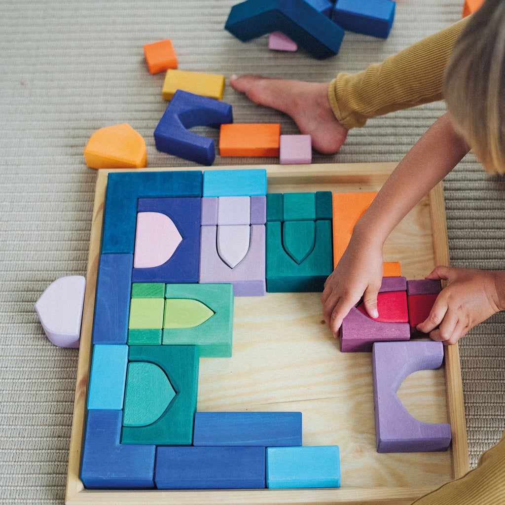 A child's hands and bare foot are shown interacting with a wooden block puzzle tray on a woven rug. The set features colorful, geometric wooden blocks in shades of blue, green, purple, orange, and pink, arranged inside a light wood frame.