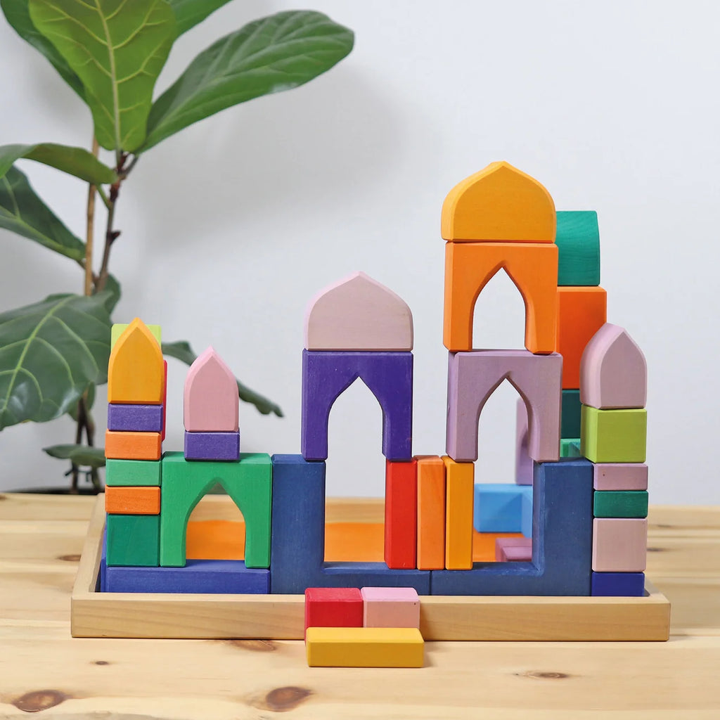 A colorful wooden building block set arranged in a castle-like structure on a light wood tray, placed on a wooden table with a green plant in the background.