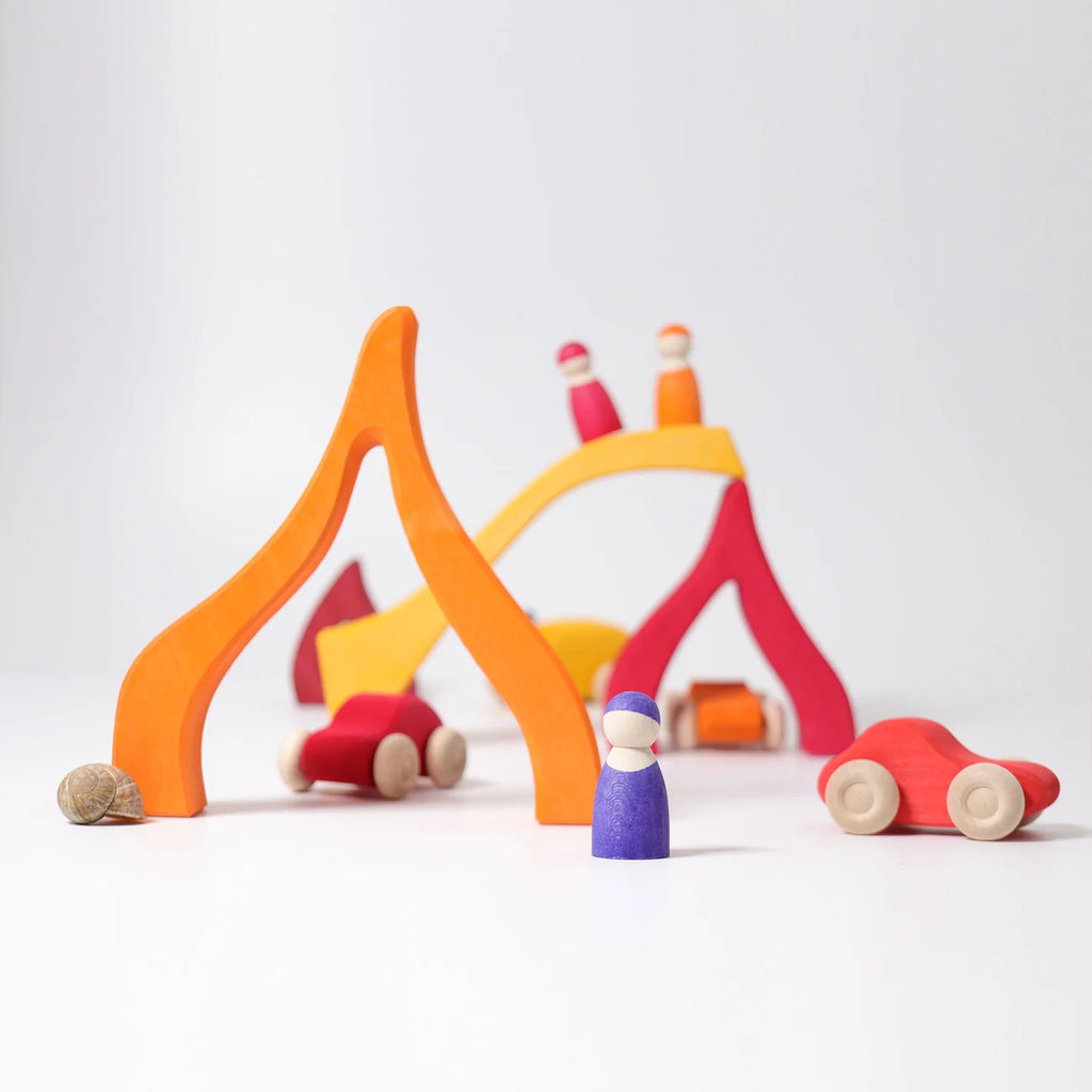 A colorful wooden toy set on a clean white background, featuring a campfire scene with orange and red logs, a yellow bridge, small red cars, and a purple peg doll.