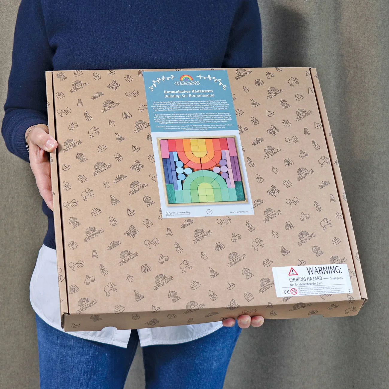 Person holding a cardboard box with a colorful product image on the label against a neutral background