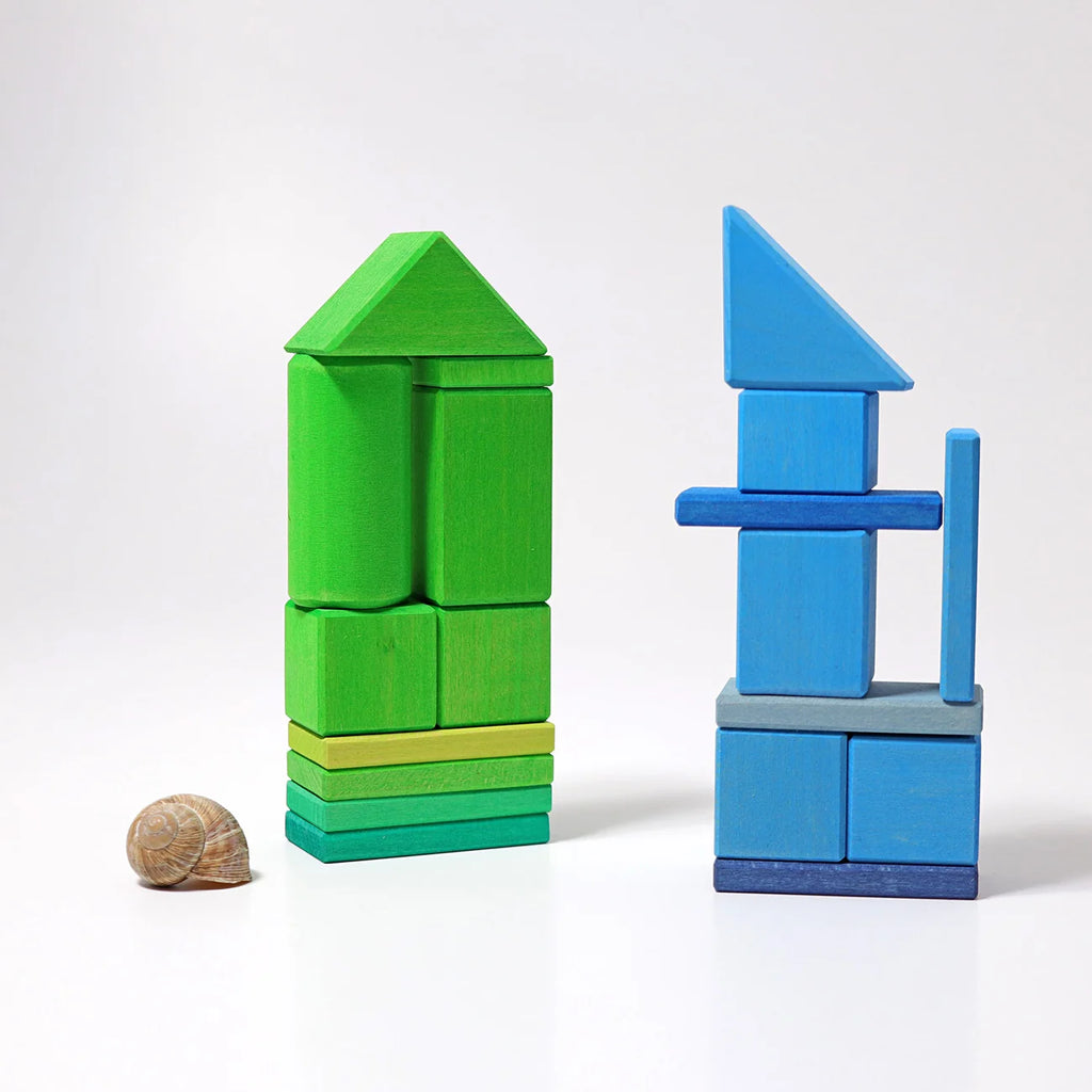 Two wooden building blocks made from painted geometric shapes, one green and one blue, displayed on a white background with a small seashell prop.