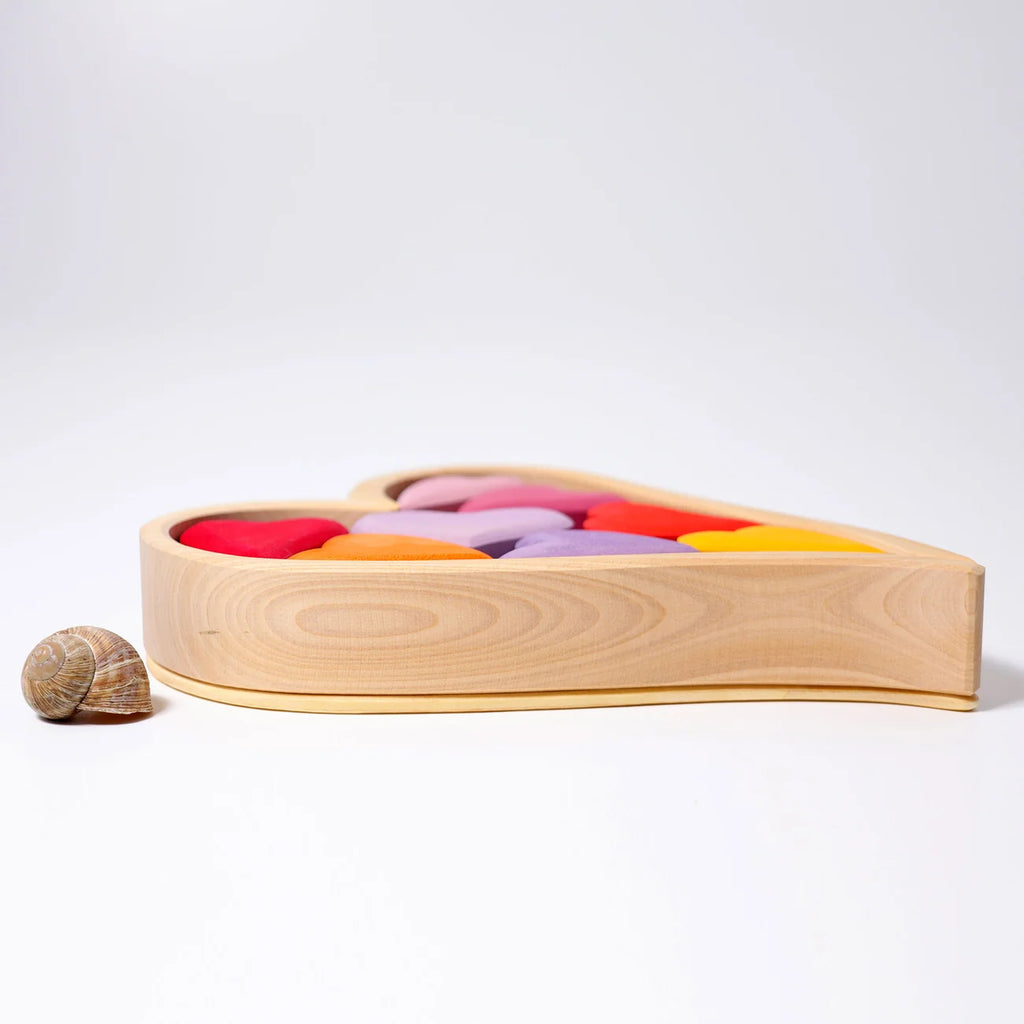 A heart-shaped wooden toy box filled with colorful smooth stones, photographed on a clean white studio background. A small snail shell sits beside the box.