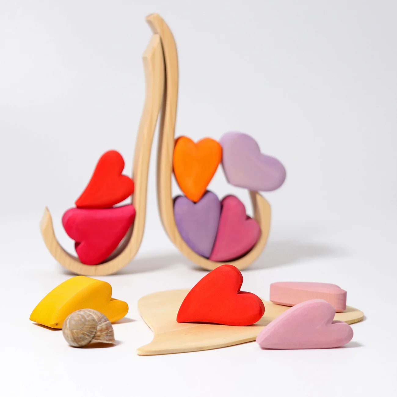 A wooden heart puzzle toy set with colorful heart-shaped pieces and a natural wood stand, displayed on a clean white background with a small seashell prop.