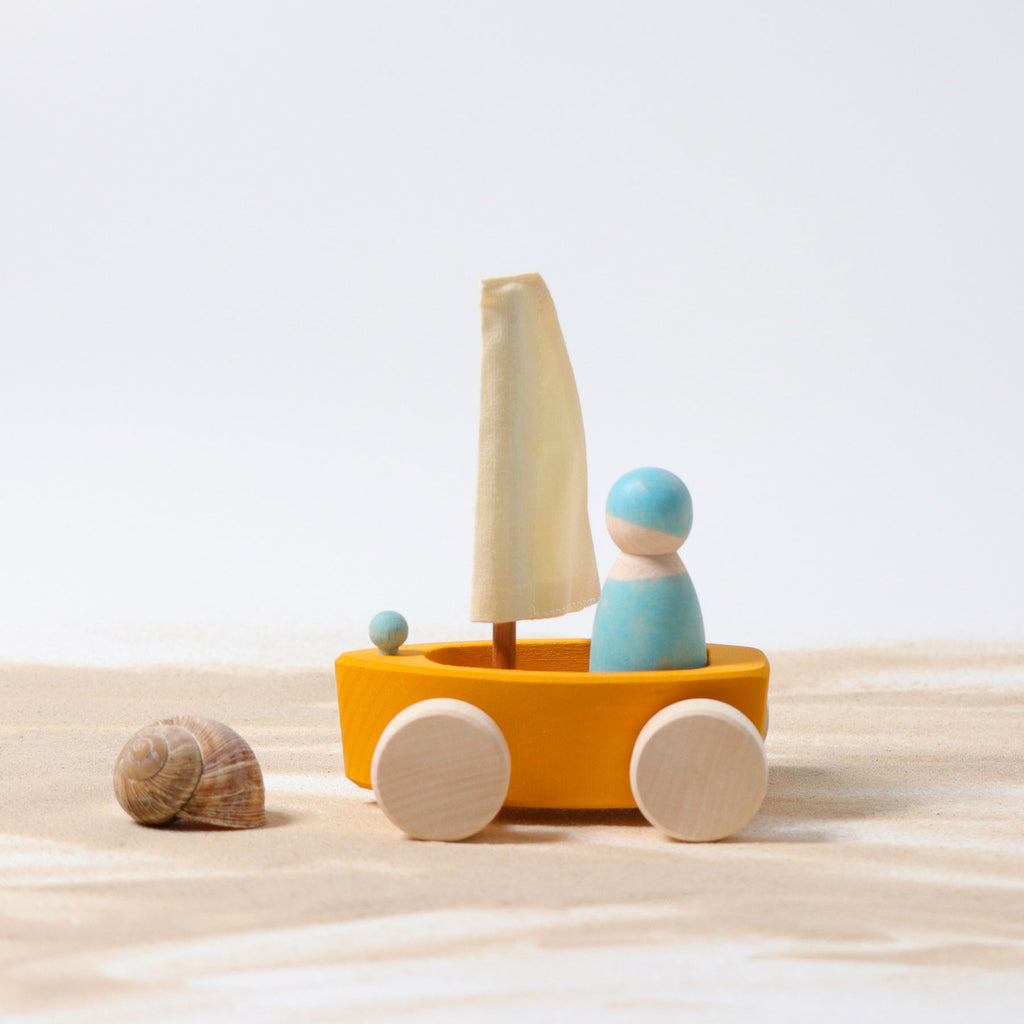 Wooden toy sailboat with a yellow body, white sail, and blue figures on a sandy surface.