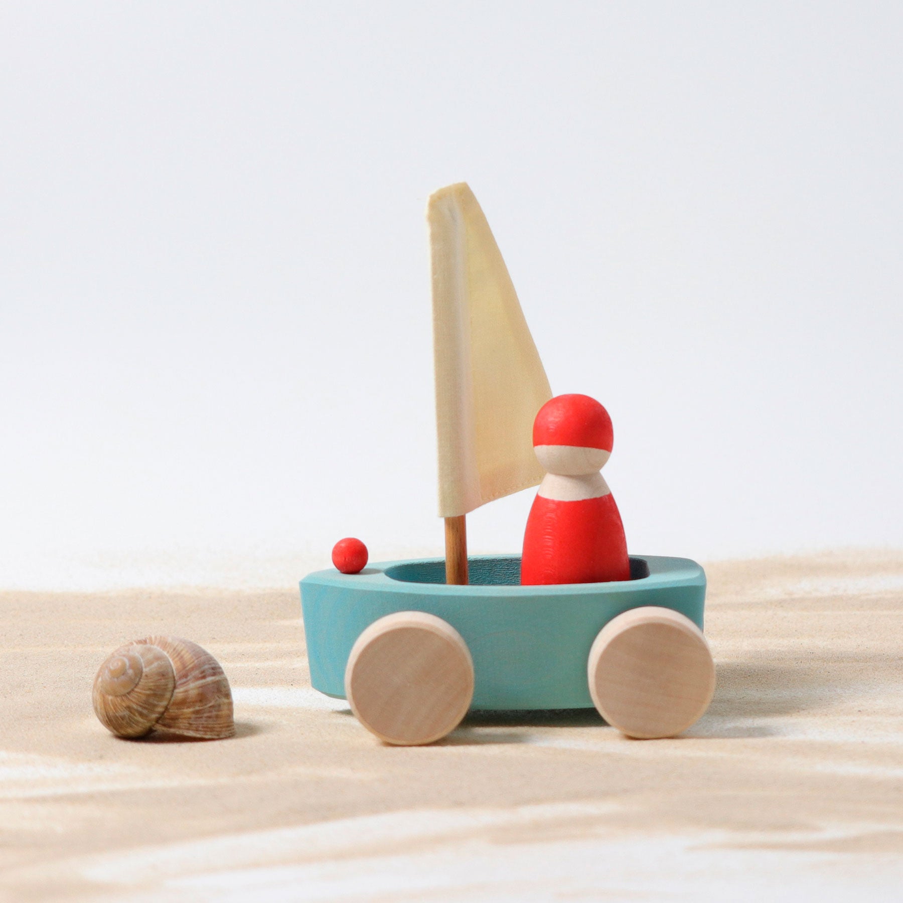 Wooden toy sailboat with a red figure on wheels, next to a shell on a light wooden surface.