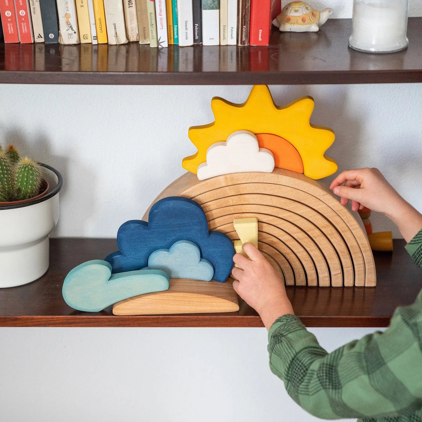 Child playing with a wooden rainbow toy on a shelf with books and a plant.