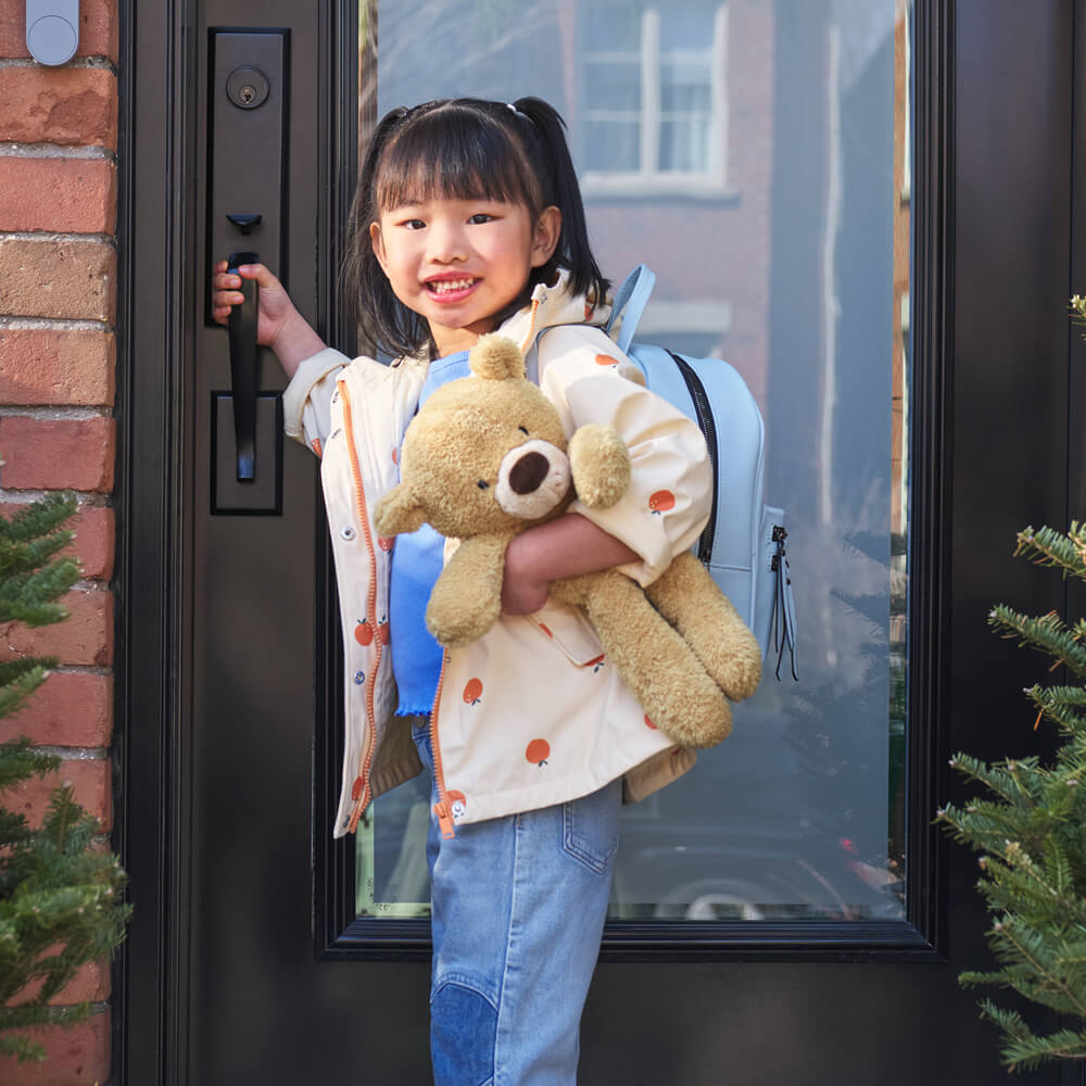 Child holding a teddy bear and a backpack, standing at a door.