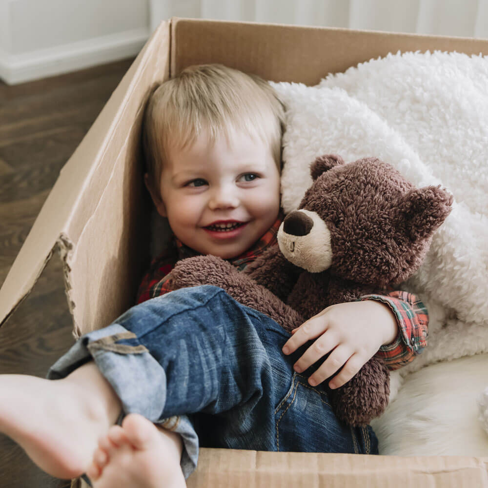 Child sitting in a cardboard box holding a teddy bear, smiling.