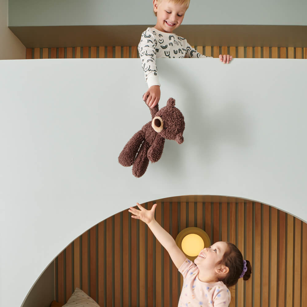 Two children playing with a teddy bear in a room with wooden floors and white walls.