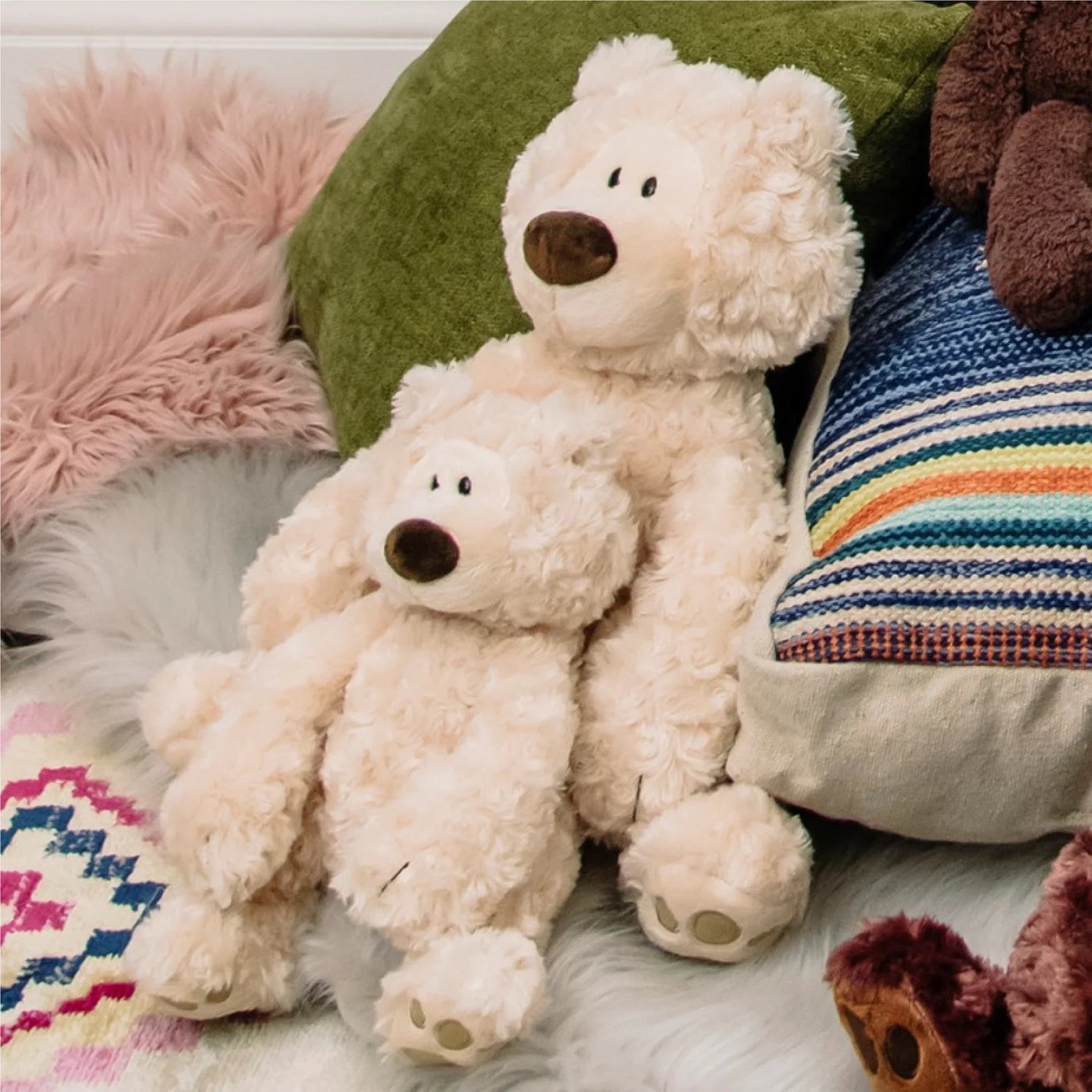 Two cream-colored teddy bears sitting on a surface with colorful pillows and blankets in the background.