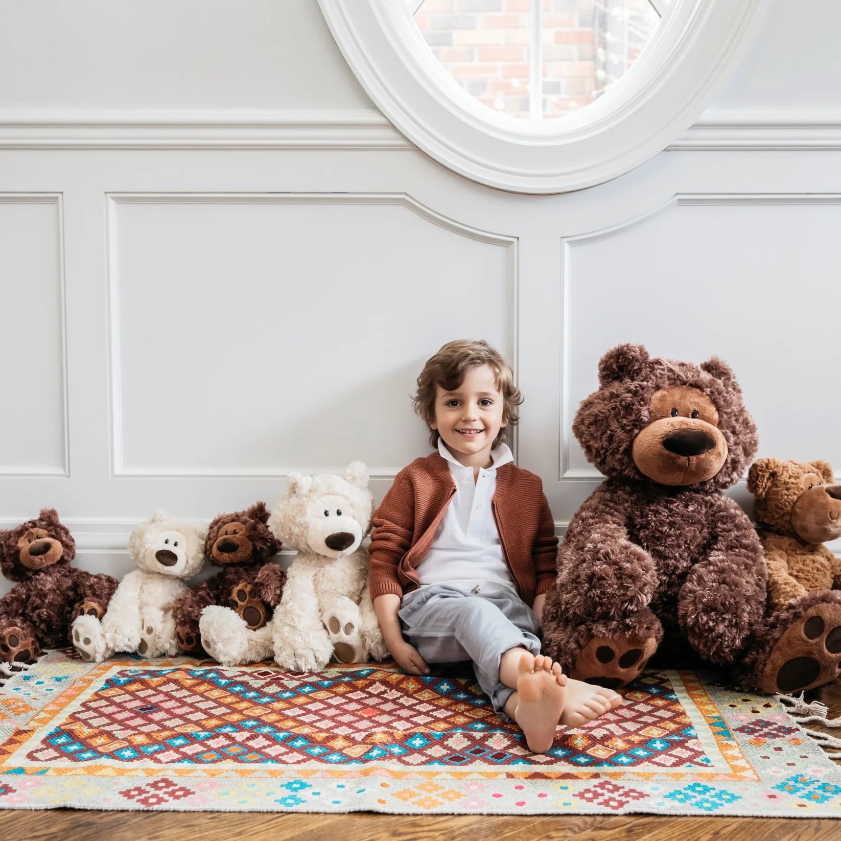 Child sitting on a colorful rug with teddy bears in a room with white paneled walls.