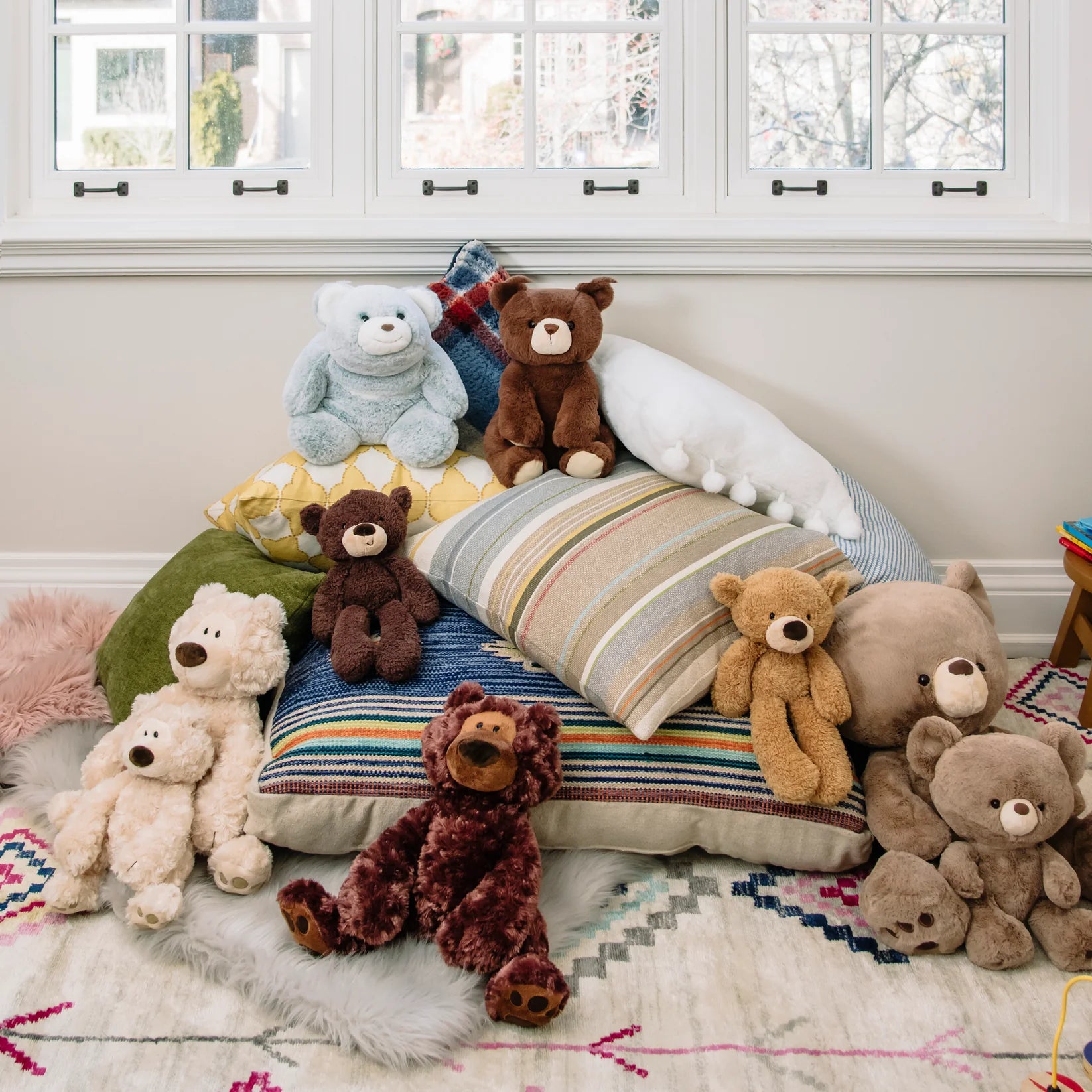 Collection of teddy bears and cushions on a bed in a room with windows.