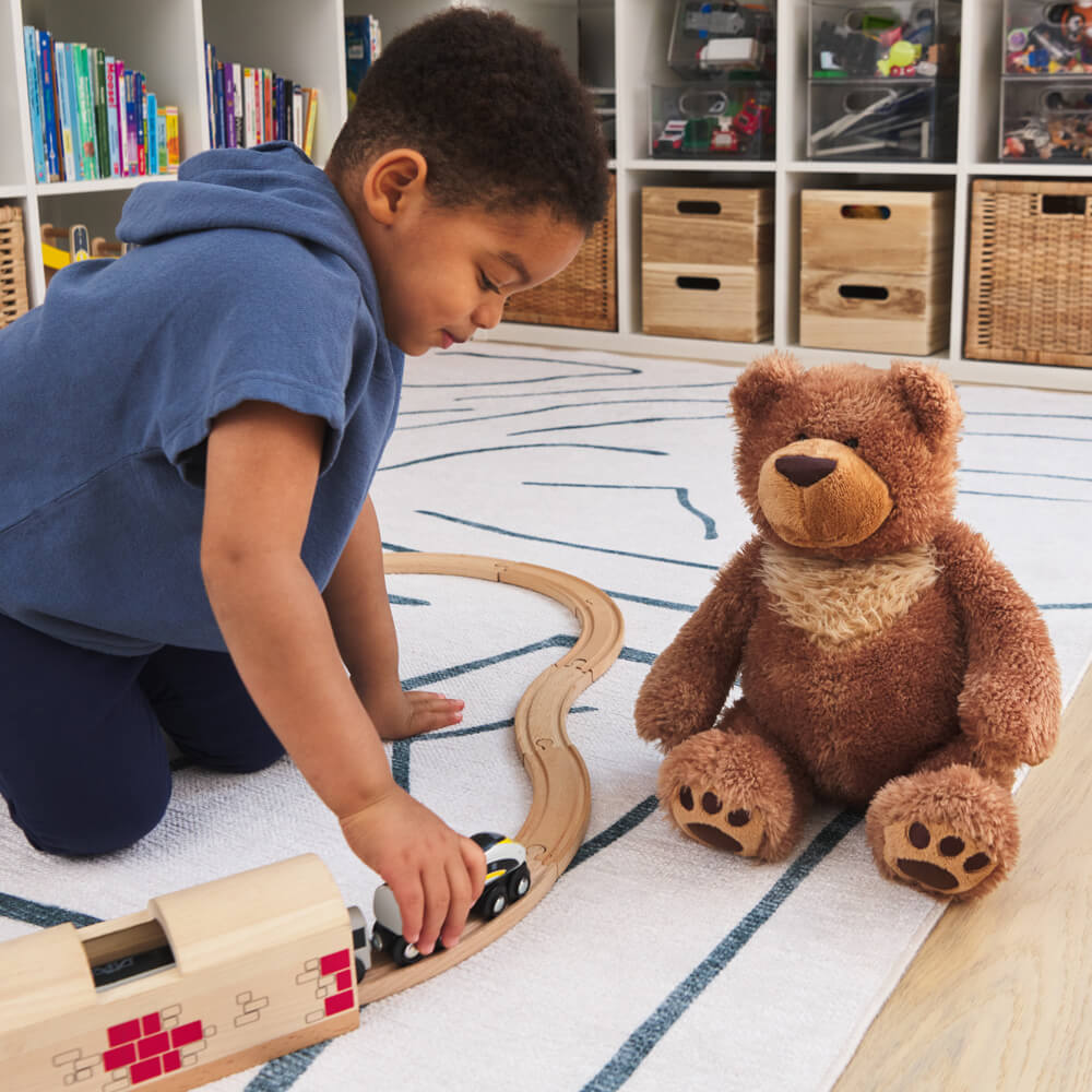 Child playing with toy train set on a rug with a teddy bear nearby