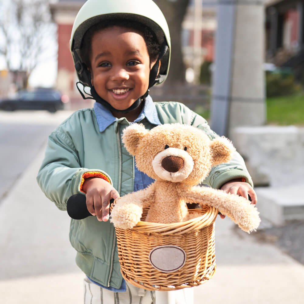 Child wearing a helmet holding a teddy bear in a bike basket on a sidewalk.