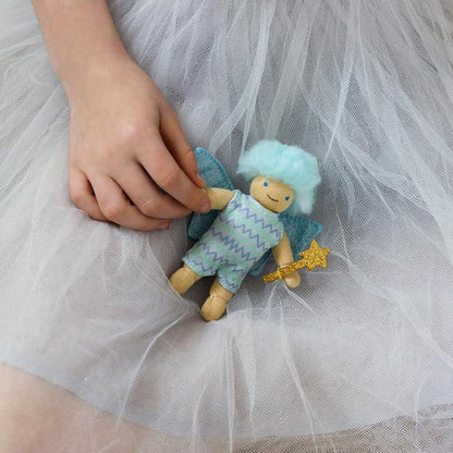 Hand holding a small fairy doll with blue hair and wings, wearing a green outfit, against a white tulle background.