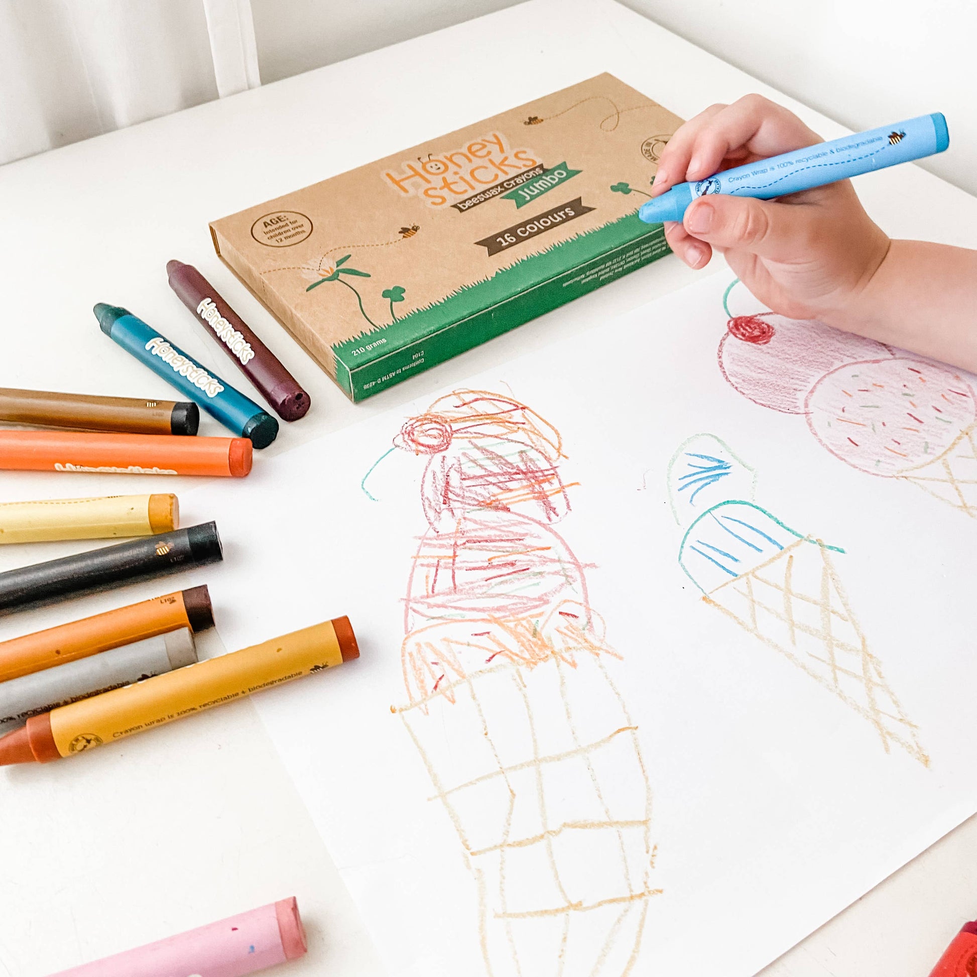 Children's hand holding a blue crayon near a box of crayons with colorful drawings on paper.