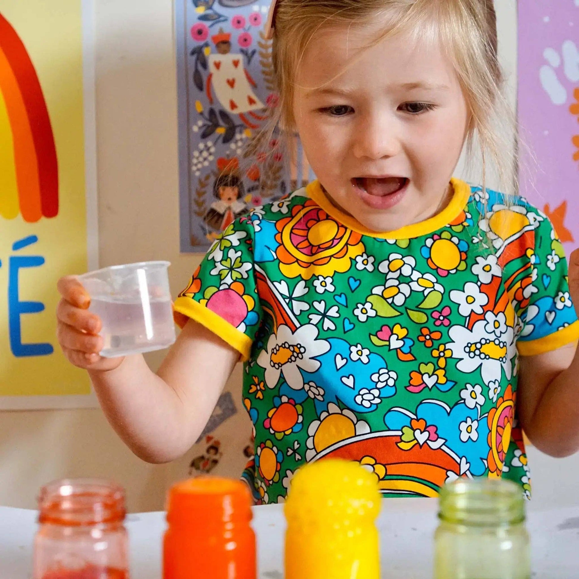 Child in a colorful shirt holding a cup with jars on a table in the background
