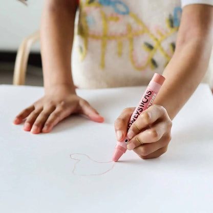 Child's hand holding a pink crayon drawing on a white surface with a colorful cushion in the background.