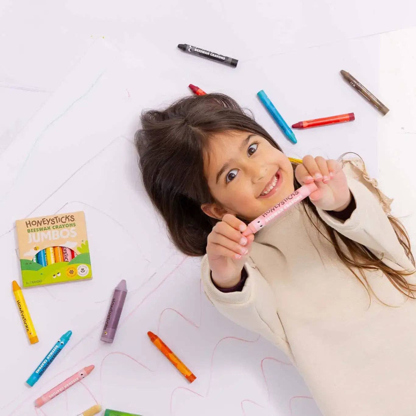 Child holding a pink crayon with colorful crayons scattered around on a white floor.