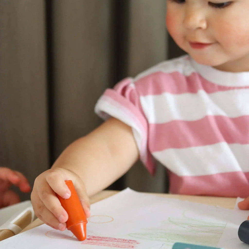 Child holding an orange crayon over a piece of paper with drawings.