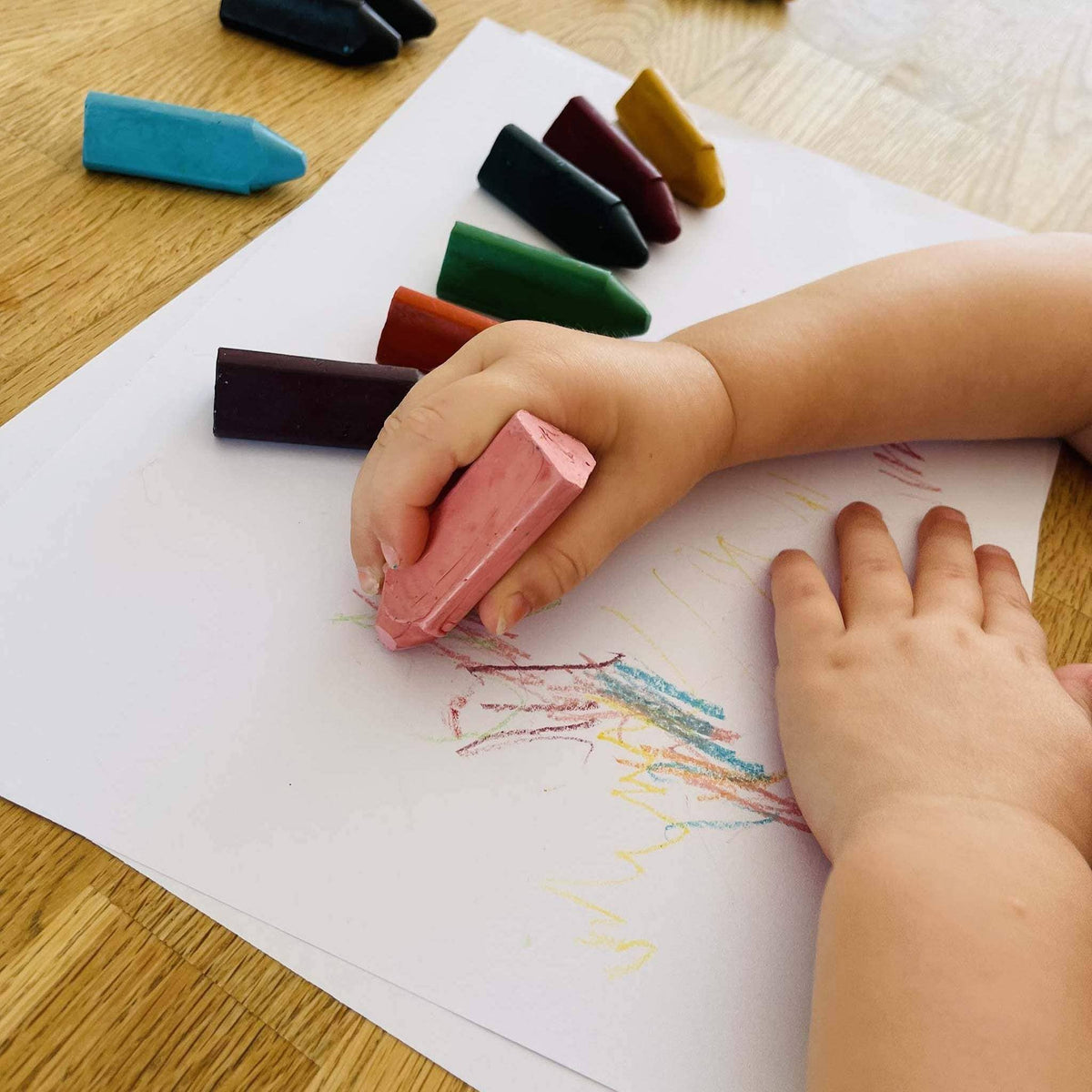 Child's hand holding a pink crayon over a drawing on white paper with crayons scattered around.