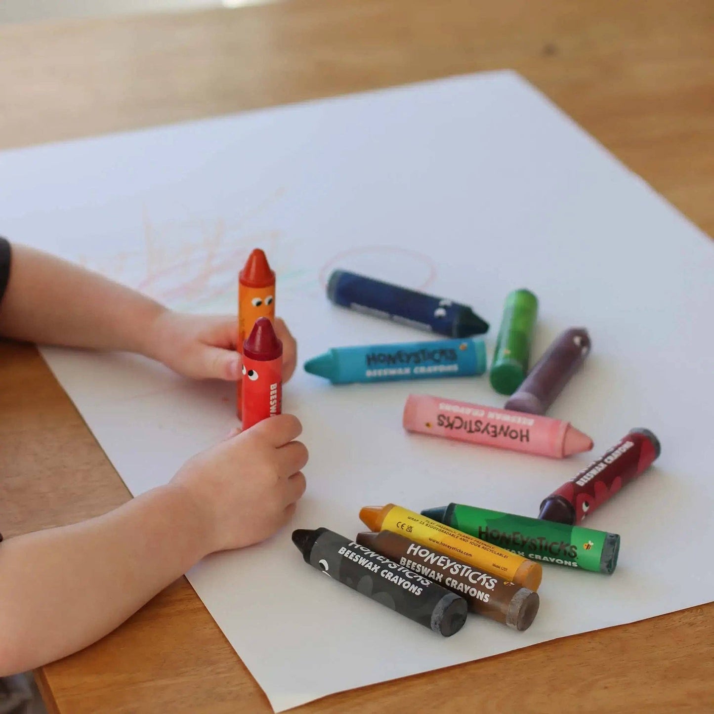 Child holding a red crayon with other crayons on a white sheet of paper on a wooden table.