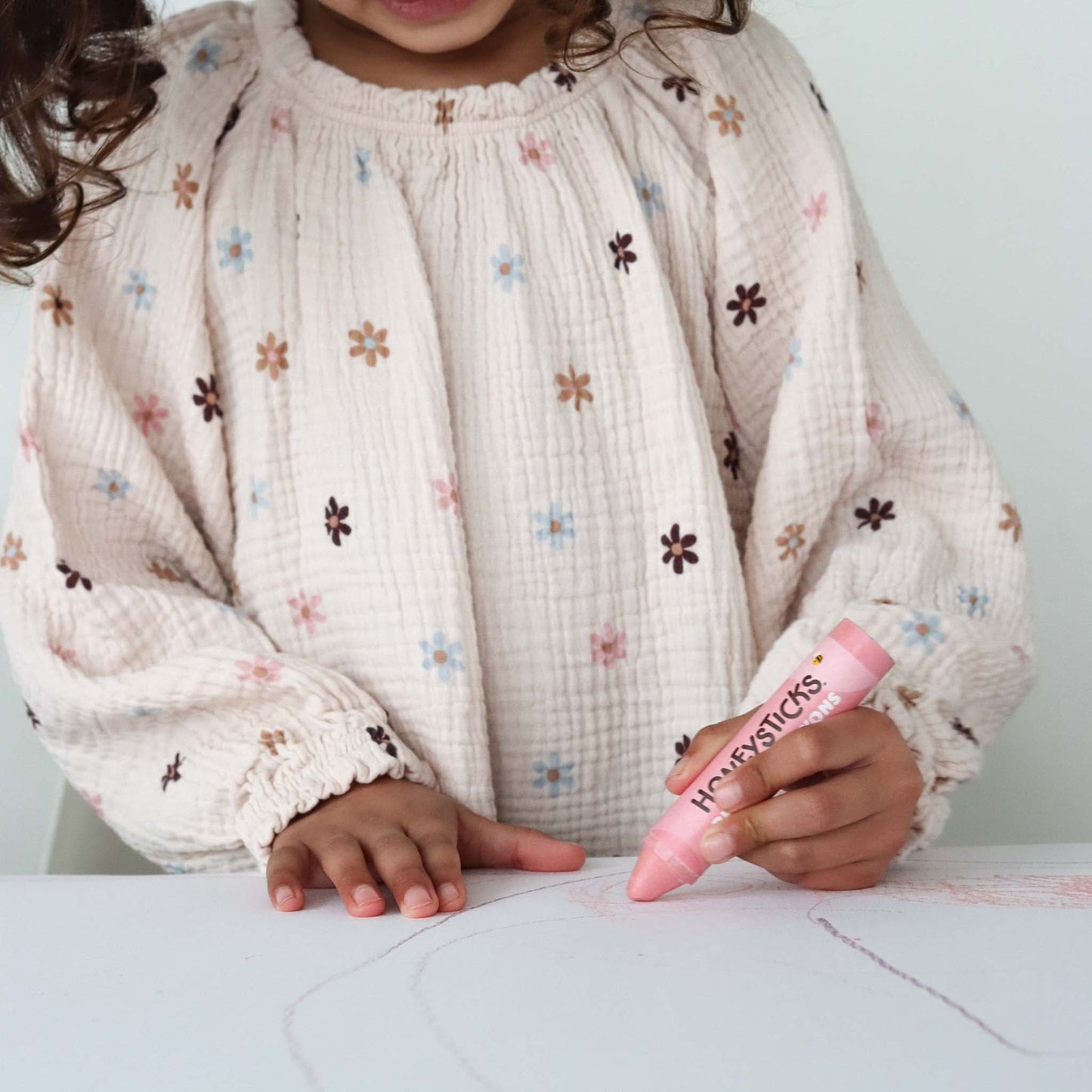 Child holding a pink crayon with 'Happiesticks' branding, wearing a floral-patterned shirt.