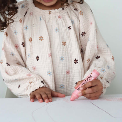 Child holding a pink crayon with 'Happiesticks' branding, wearing a floral-patterned shirt.