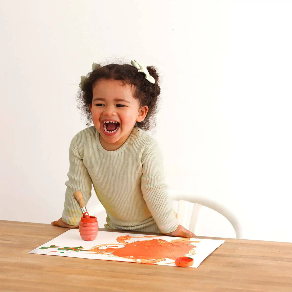 Child painting on a paper with a brush and paint container, sitting at a table.
