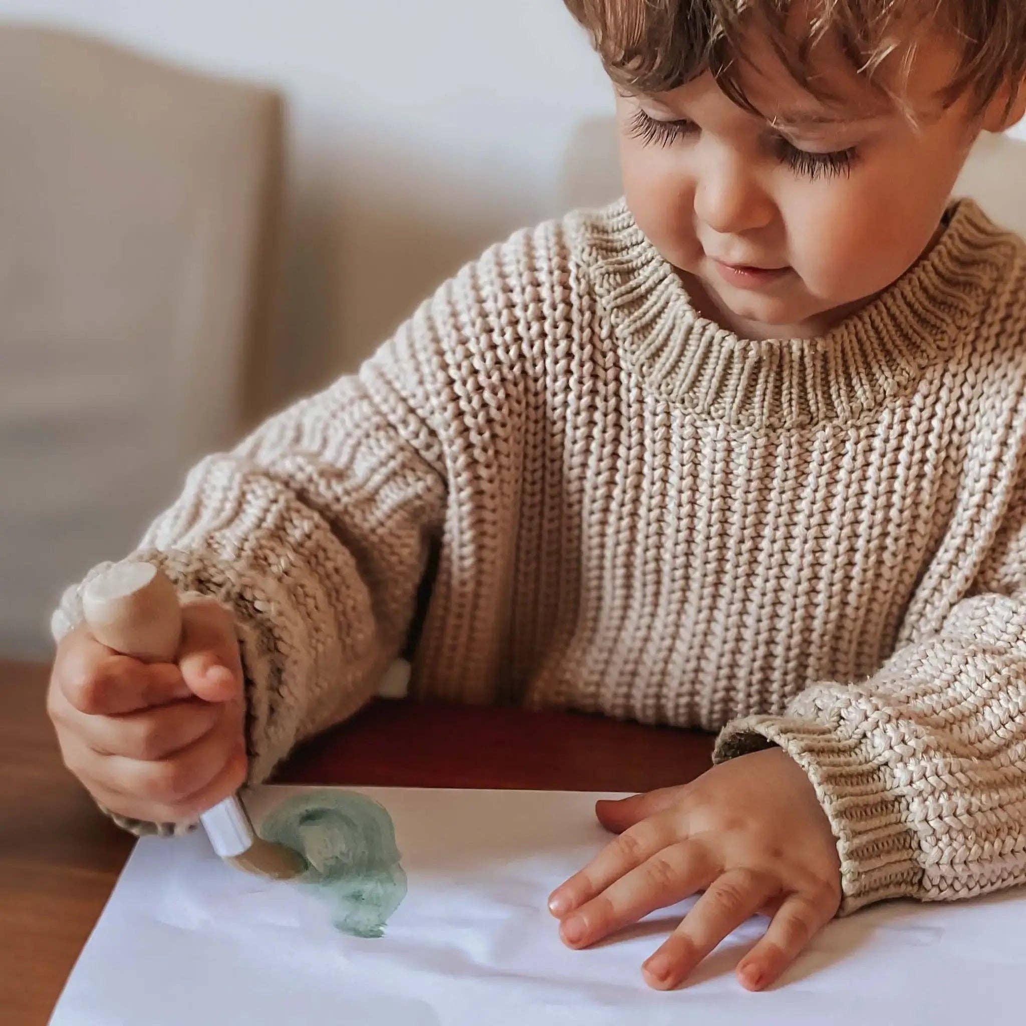 Child in a beige sweater painting on a white surface.