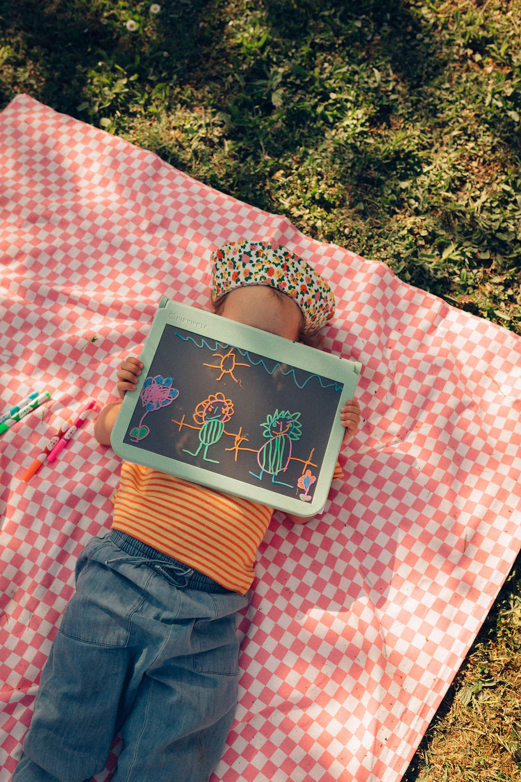 Child lying on a pink checkered blanket outdoors, holding a tablet with colorful drawings.