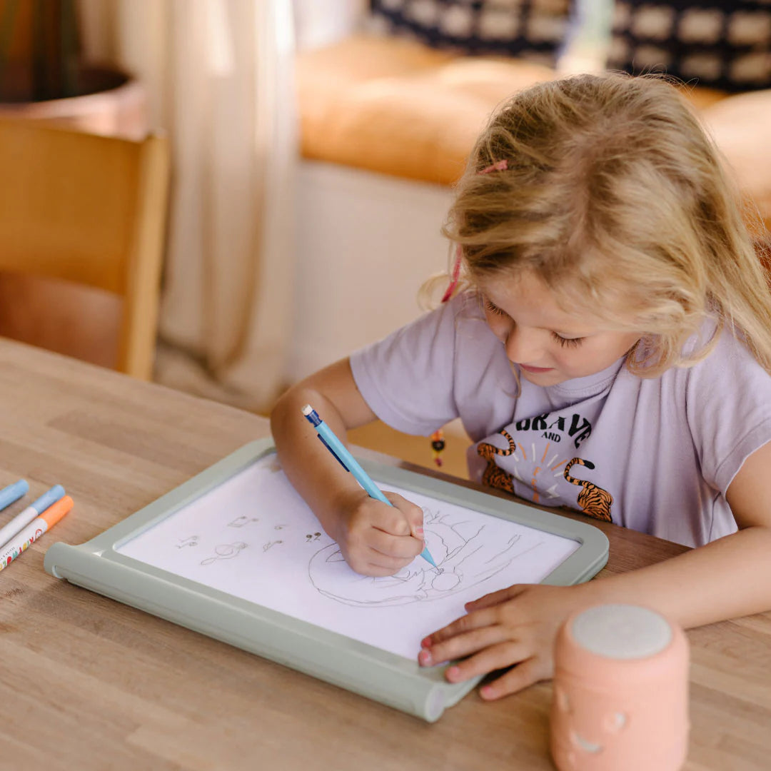 Child drawing on a tablet with markers at a wooden table.