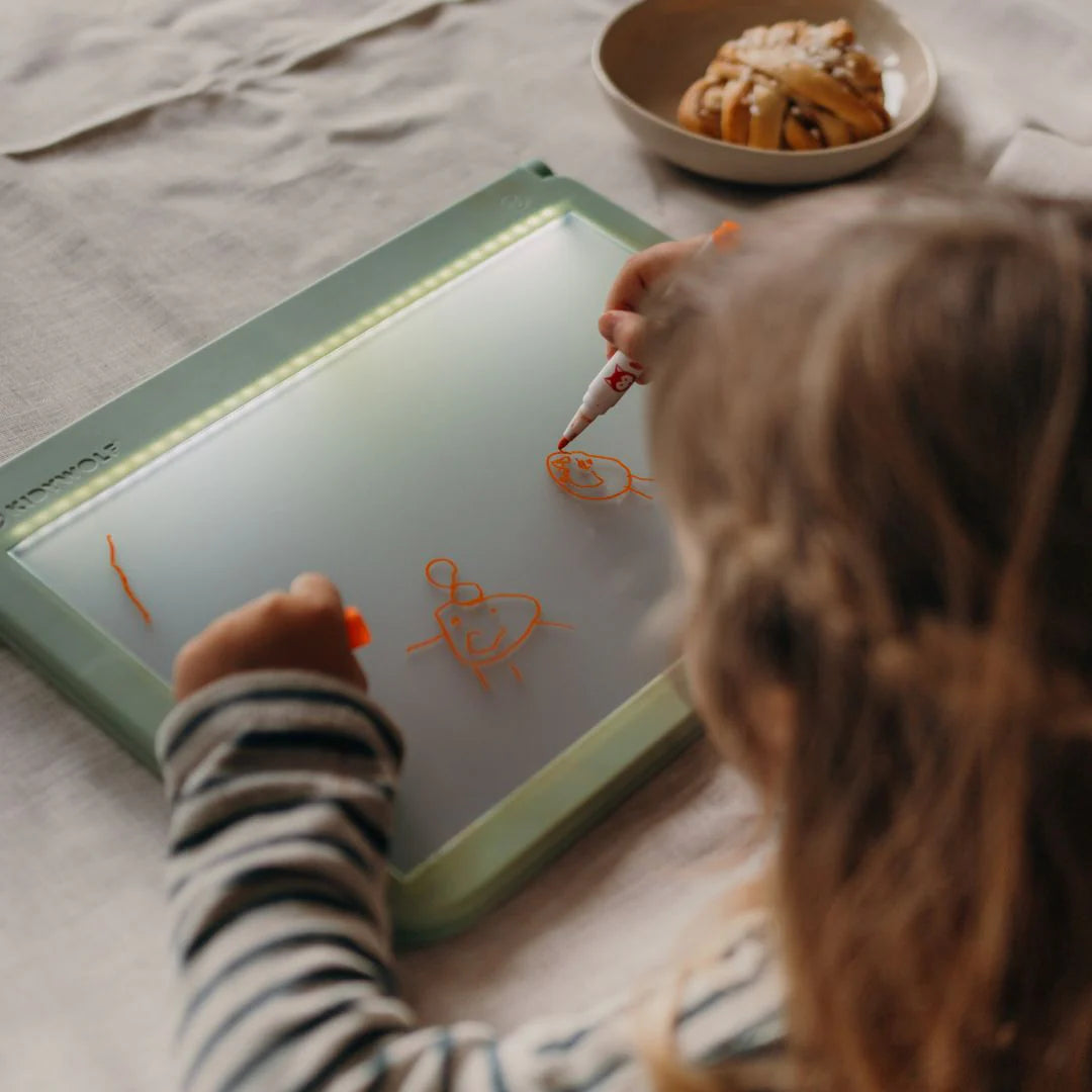 Child drawing on a light-up board with orange markers, sitting at a table.