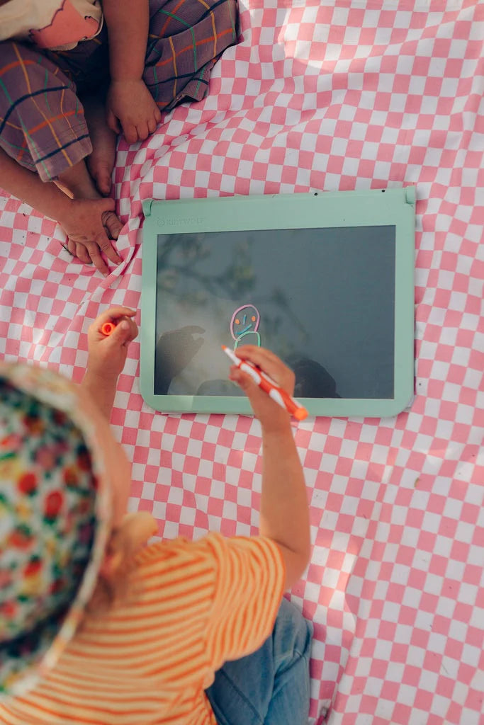 Child drawing on a digital tablet with a stylus on a pink checkered blanket.
