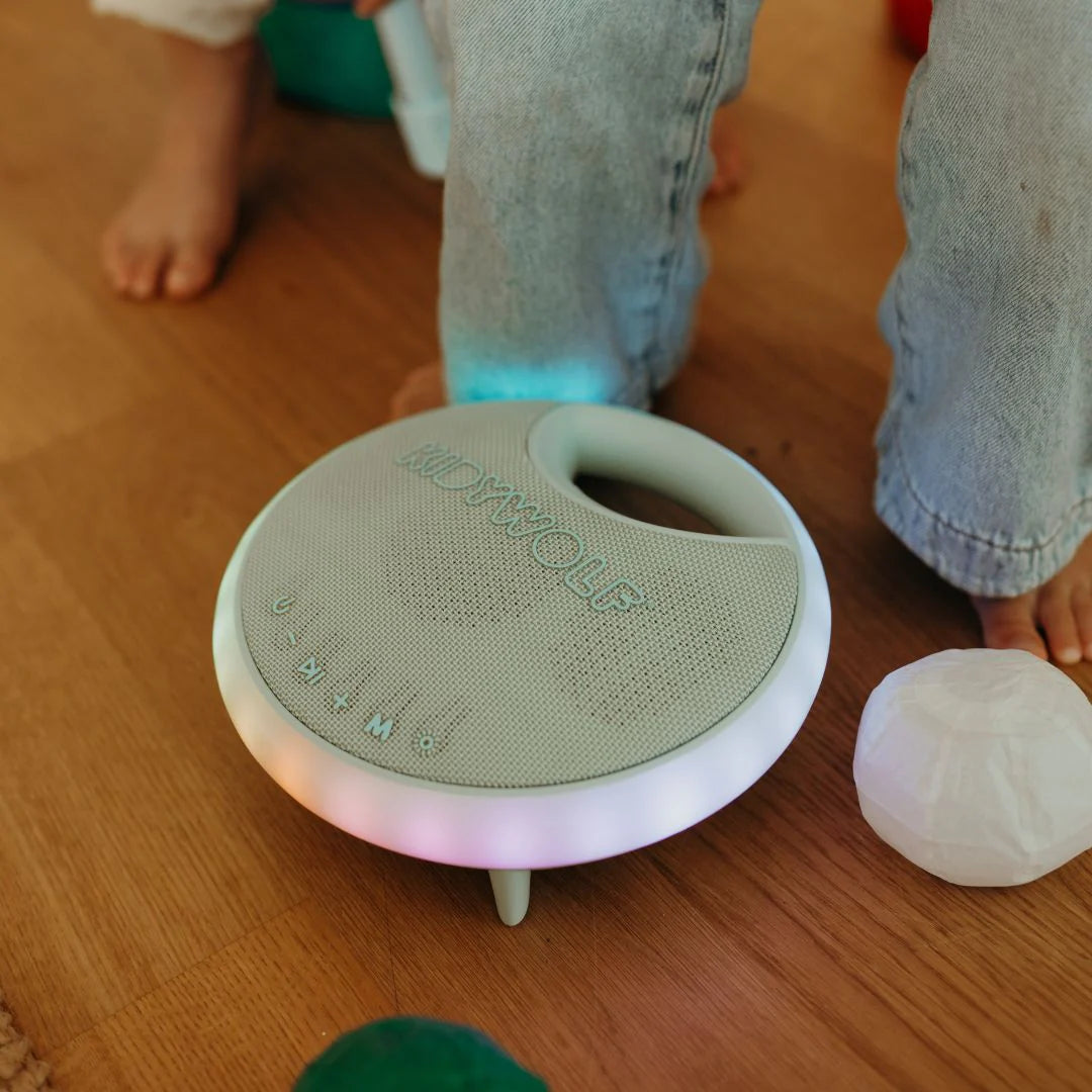 Round electronic device with glowing base on a wooden floor, surrounded by children's feet and toys.