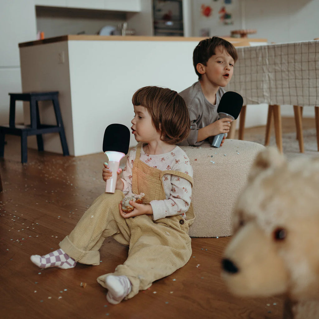 Two children sitting on a couch holding black and white toy microphones in a room with a teddy bear in the foreground.