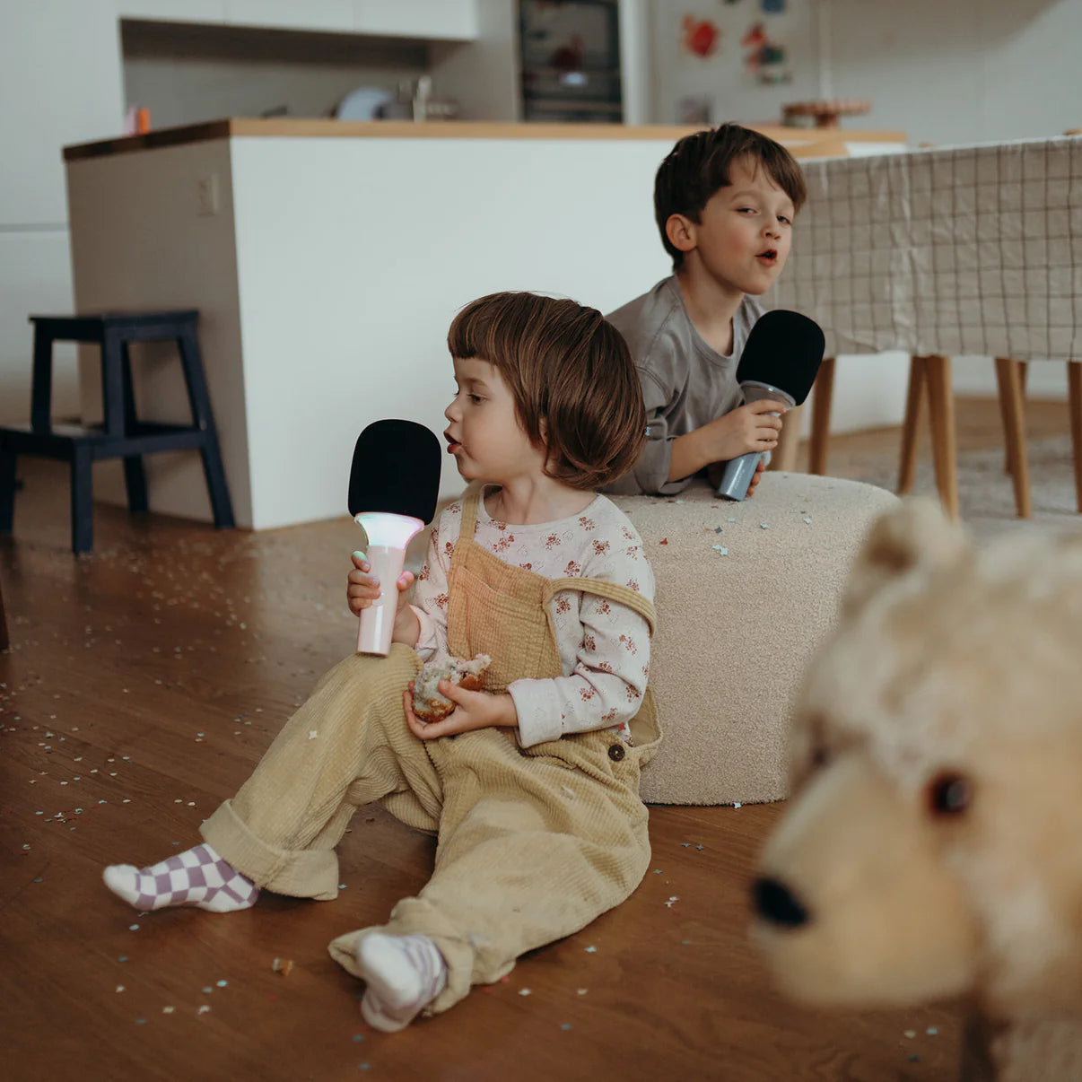 Two children sitting on a couch holding black and white toy microphones in a room with a teddy bear in the foreground.