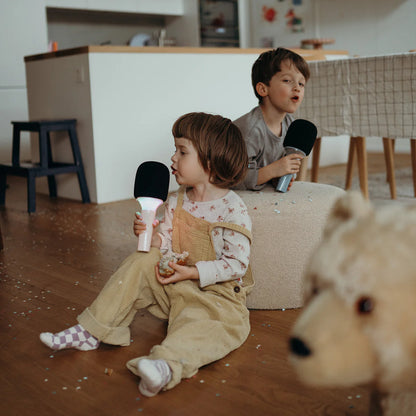 Two children sitting on a couch holding black and white toy microphones in a room with a teddy bear in the foreground.