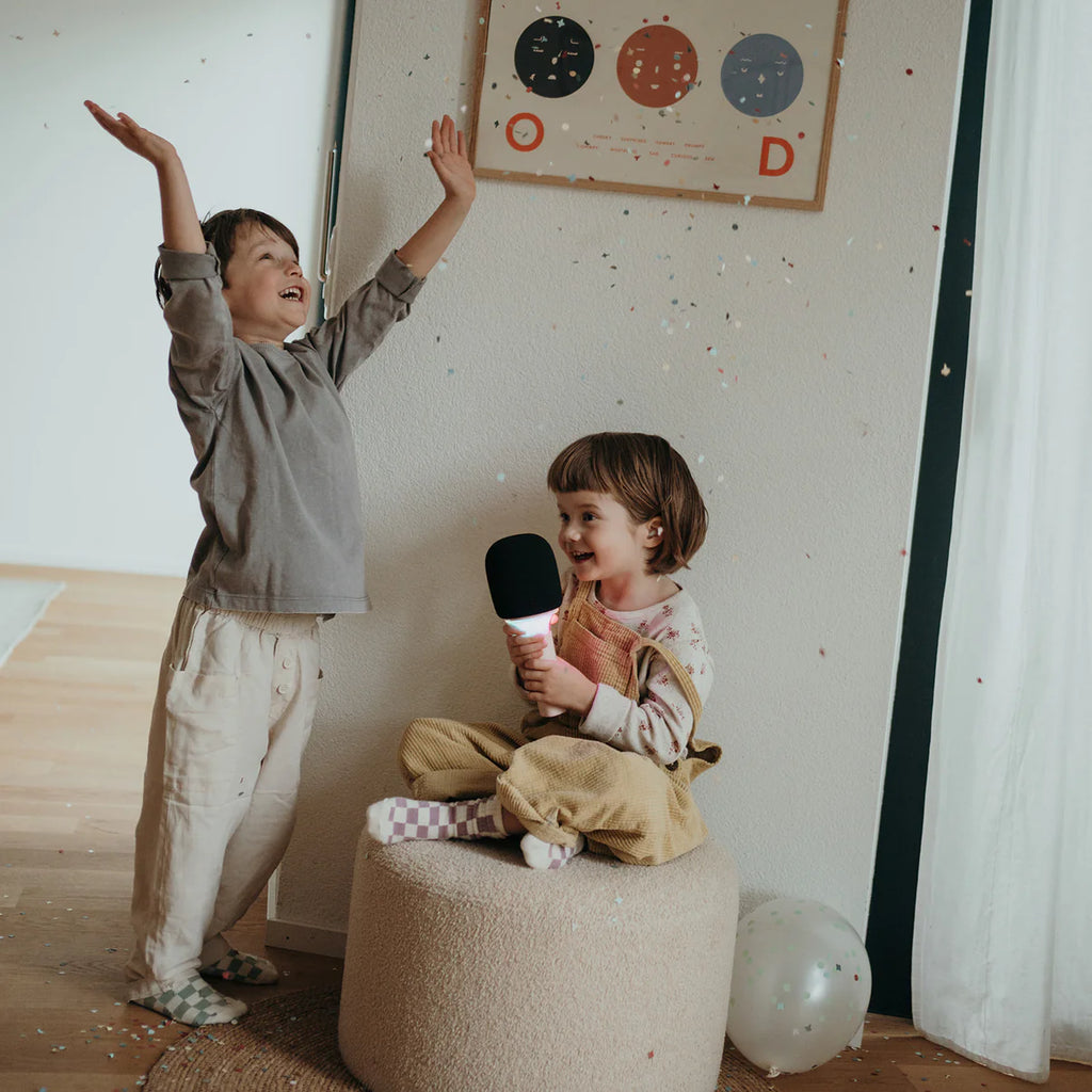 Two children in a room with a child sitting on a stool holding a microphone and another child standing with arms raised.