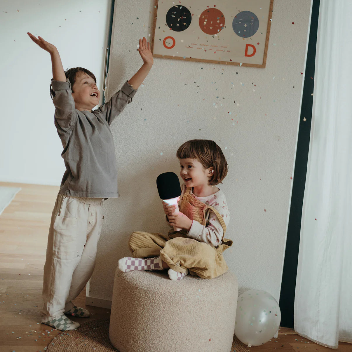 Two children in a room with a child sitting on a stool holding a microphone and another child standing with arms raised.