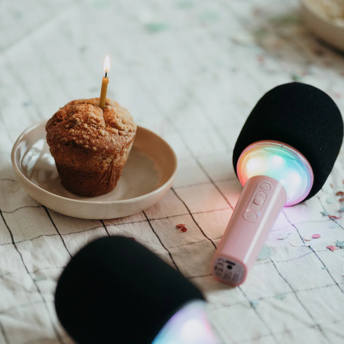 Muffin with a lit candle on a plate next to a microphone with colorful lights on a patterned surface.