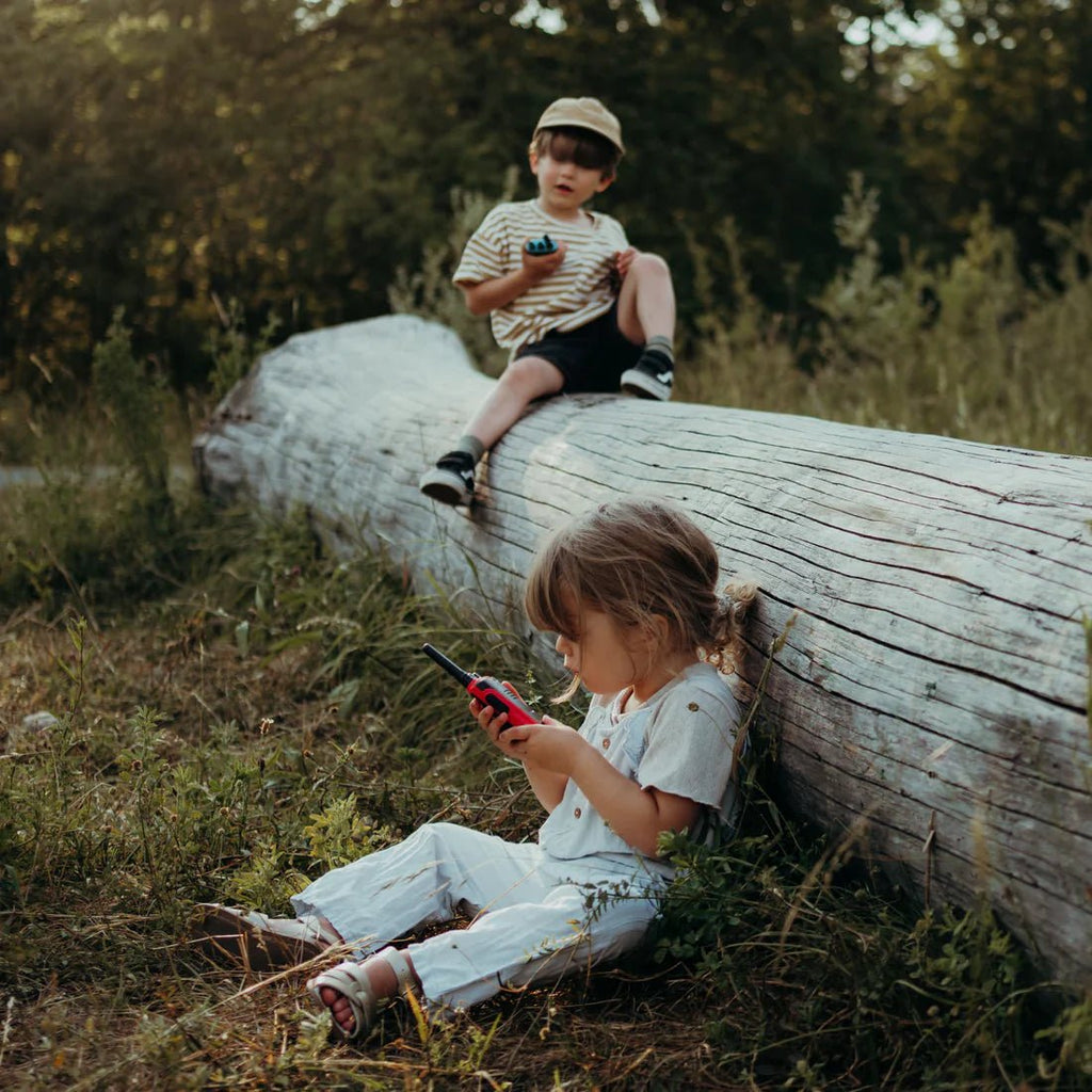 Two children sitting on a log in a natural setting, using electronic devices.